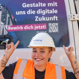 Eine junge Frau mit Wahnweste und einem weißen Helm mit dem Logo der Marke Axians lächelt und zeigt mit beiden Zeigefingern nach oben. Über ihrem Kopf steht der Text: „Gestalte mit uns die digitale Zukunft“ und „Bewirb dich jetzt!“.